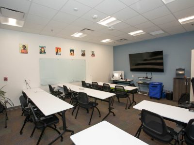 A classroom with white tables, black chairs, a large wall-mounted monitor, a whiteboard, and several small portraits on the wall.